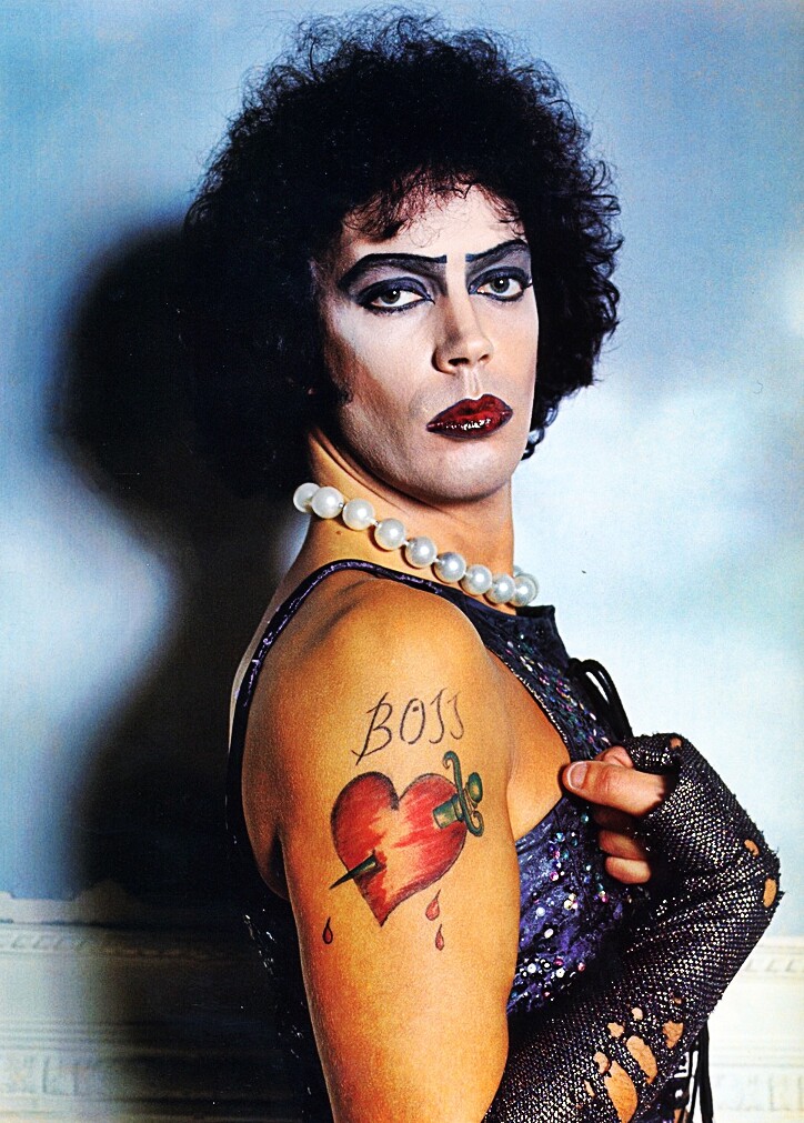 Disney Princess Dr. Frank N. Furter, played by Tim Curry. A tall, striking trans woman wearing a black negligee and a large string of pearls. Her makeup is impeccable and her shoulder has a tattoo of a sword-pierced heart labeled BOSS.
