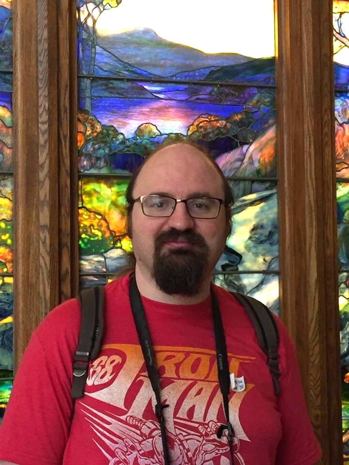 A masculine presenting person with white skin and Mediterranean features, a goatee beard, receding hair pulled back into a pony tail, and glasses, smiling gruffly. They're wearing a red "Iron Man" t-shirt, a backpack, and camera strap, and are standing in front of a beautiful stained glass window showing mountains, a lake, and trees.