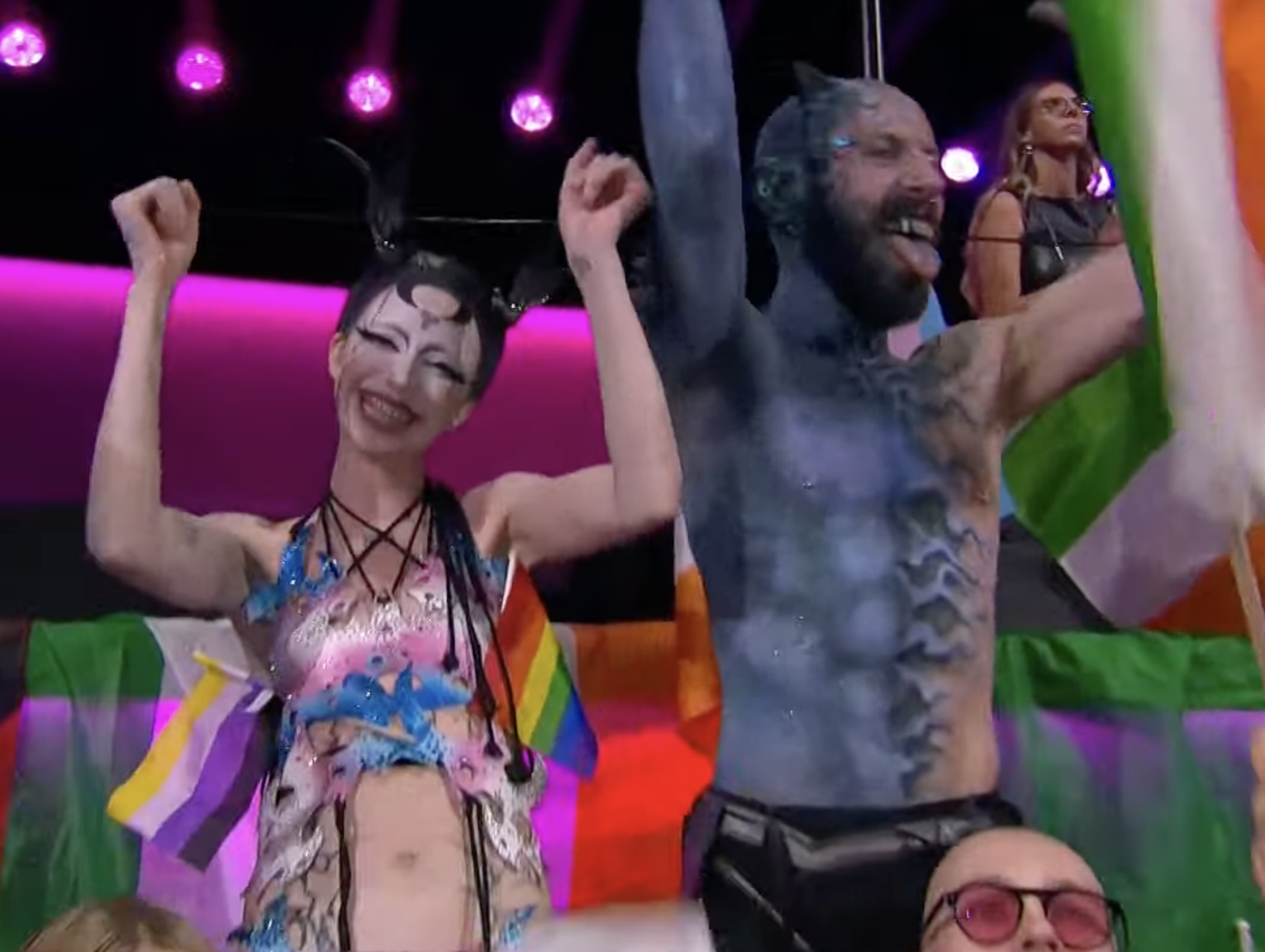 Two people in vibrant costumes, one with a horned headpiece, celebrating with raised arms, against a backdrop of multicolored flags. A woman with black horns, braces and a dress in transgender flag colors smiles and cheers. Pride flags are poking out of the dress as well.