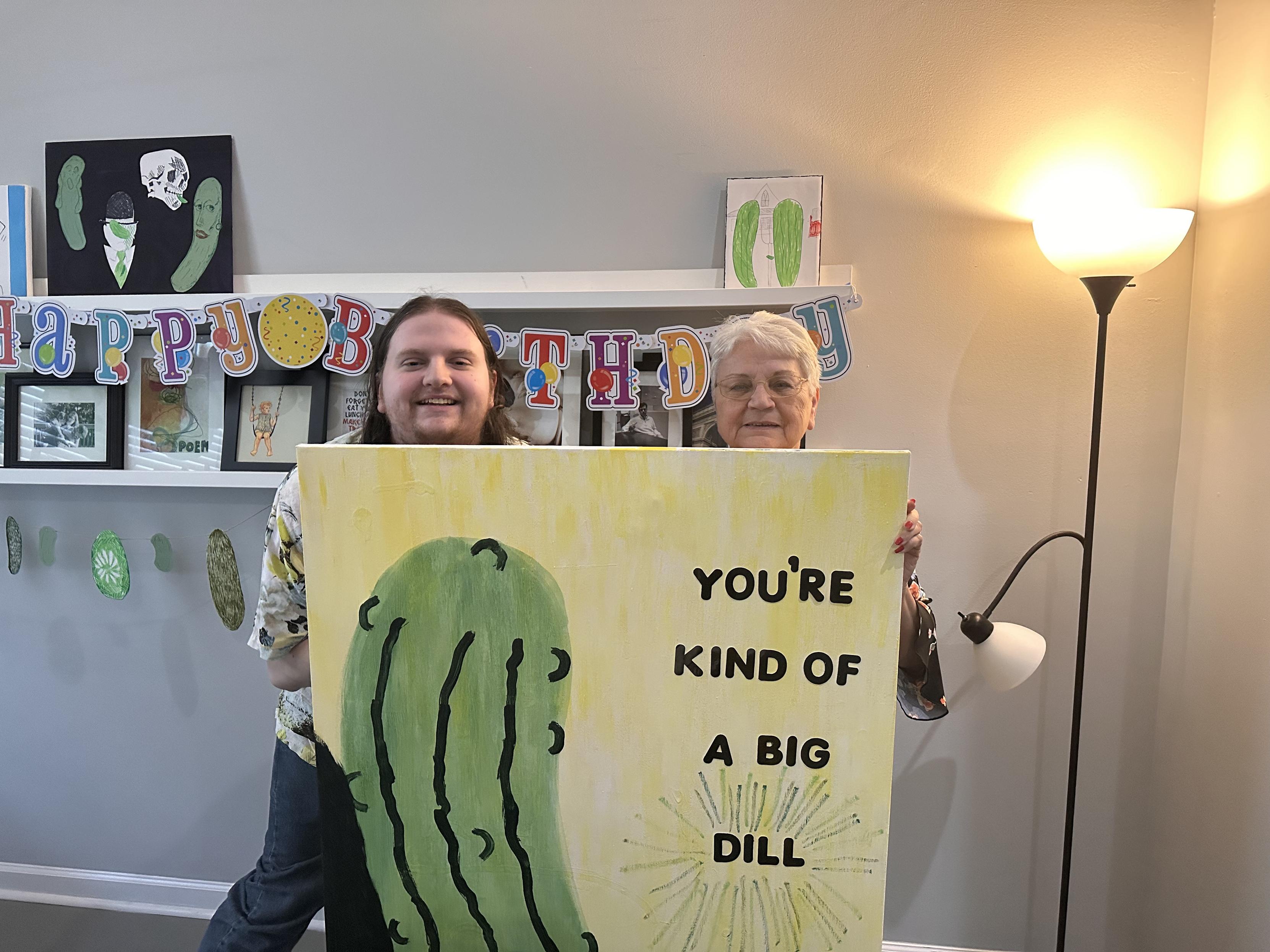 The artist and the birthday girl holding a large painting of a pickle that has the text “you’re kind of a big dill” on it. 