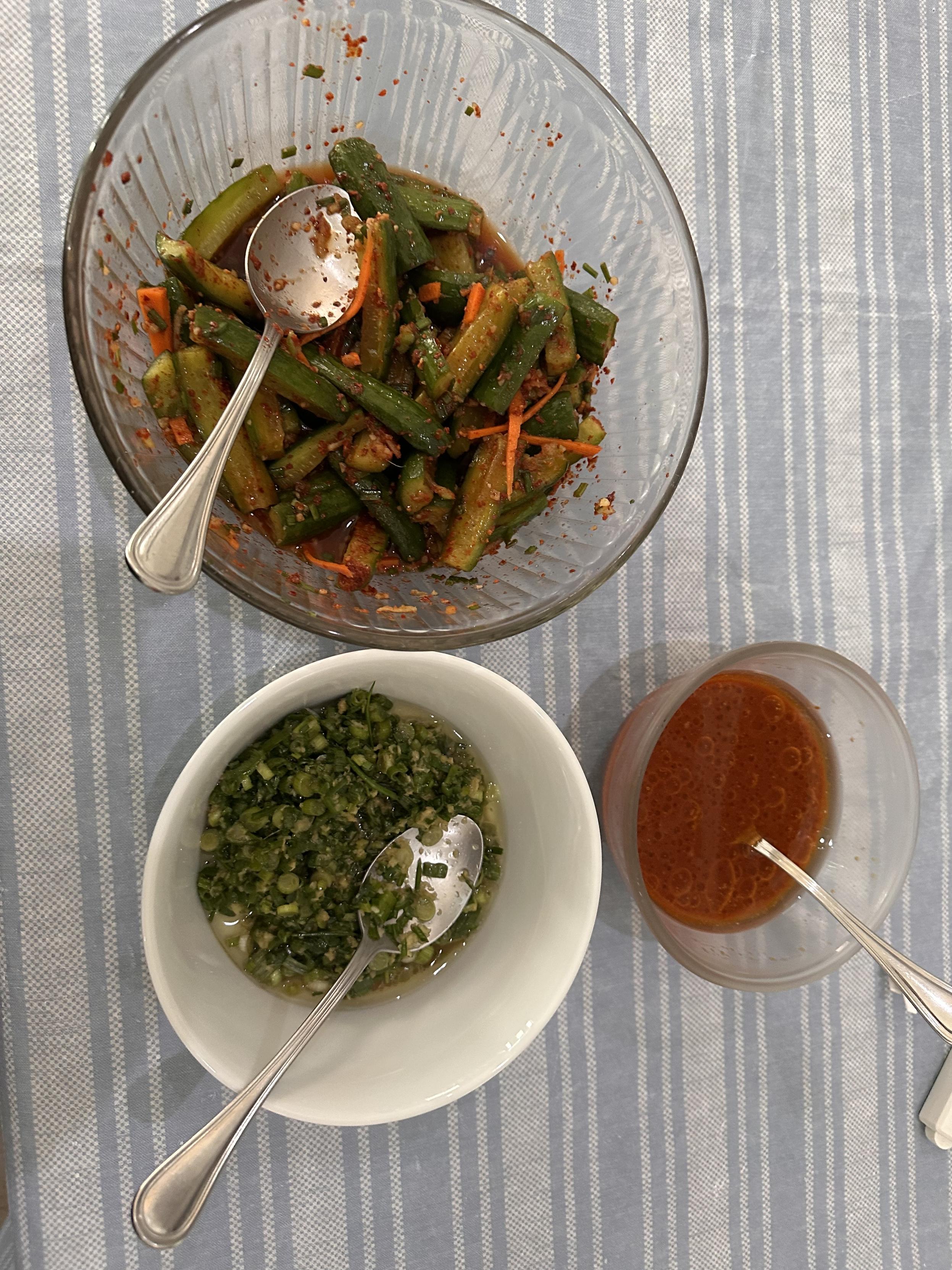 A tops down view of three bowls. One filled with cucumber kimchi (dark green cucumber sticks, red with chili, and pops of little carrot sticks), one with ginger scallion sauce (bright green scallions glistening with oil and ginger) and one with ssam sauce (it’s brown and looks a little like ketchup). 