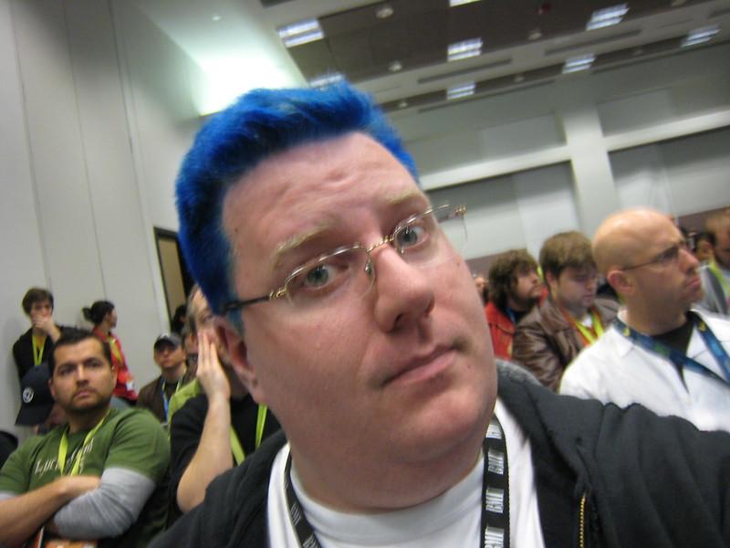 A self portrait of a man with newly dyed bright blue hair looking slightly apprehensive sitting in a brightly lit conference session wearing glasses and a hoodie.