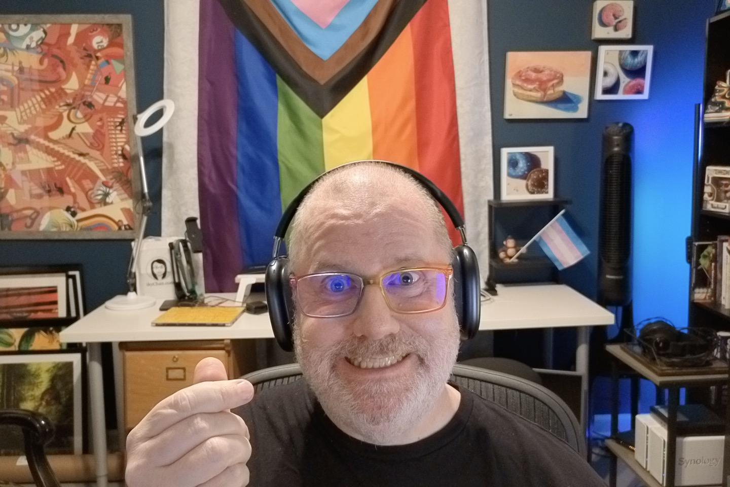 A middle-aged, balding, bearded white guy wearing rainbow glasses gives finger hearts and smiles stupidly into the camera, proudly showing you his GIGANTIC Pride flag and small Trans Pride flag in the background.