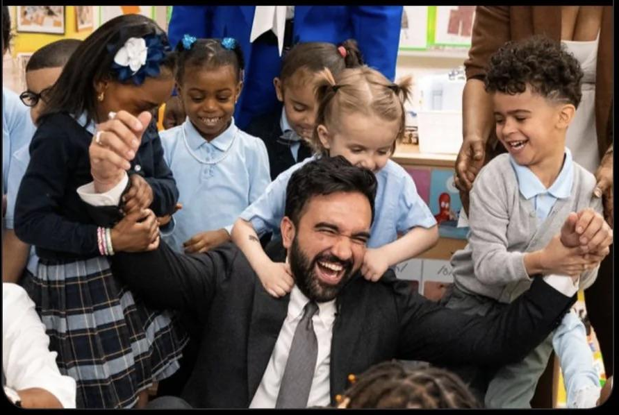 Mayor of NYC Zohran Mamdani surrounded by kids in school pretend-playing he's being overpowered. He's got a huge open-mouthed grin on his face that you could pretend is anger if you were disingenuous.