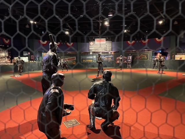 A photo of the baseball diamond exhibit at the Negro Leagues Baseball Museum.  It's a replica baseball diamond with bronze statues of men standing in position ready for the next pitch.  The photo is taken from behind the netting behind home plate.