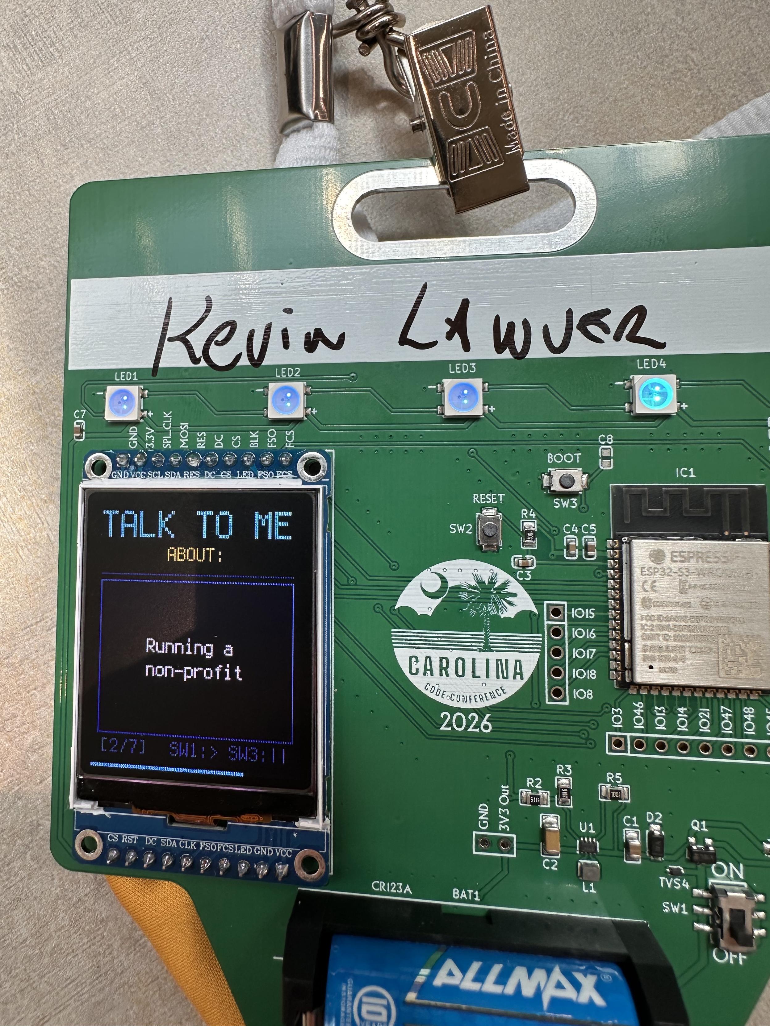 A conference badge that’s also a green circuit board. It has a small rectangular screen in the bottom left corner that says “talk to me about running a non-profit.”