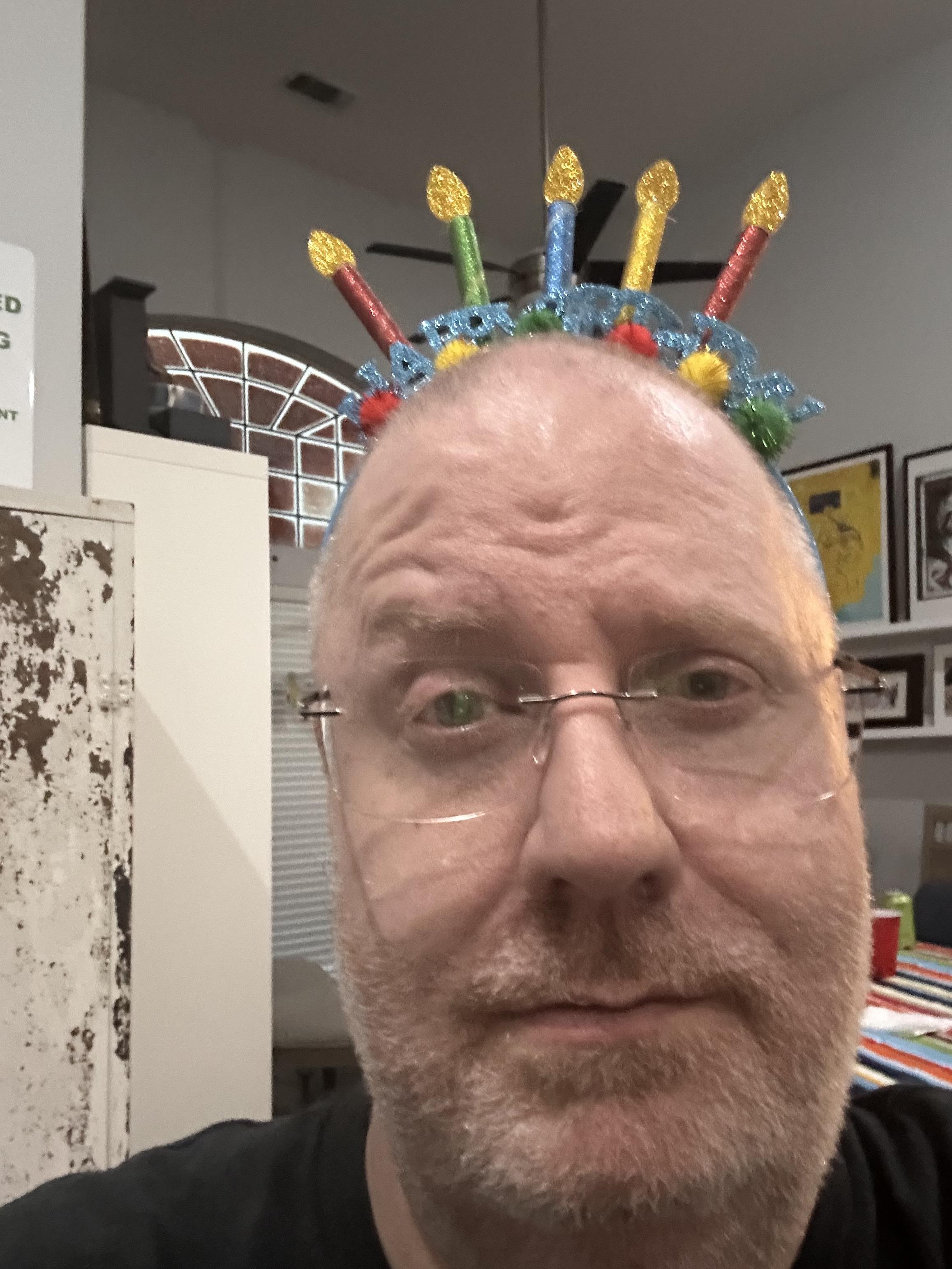 A selfie of a bald middle aged man wearing a birthday headband with a raised eyebrow. He might be a little tipsy. 
