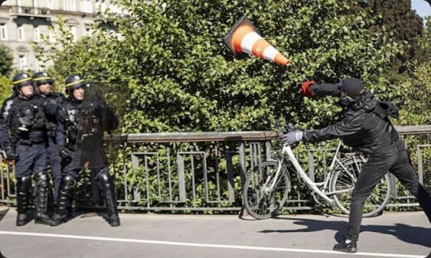 Photo from an article about the current protests in France showing a protestor on the right side of the frame and three cops on the left. the cop closest to the protestor has a riot shield up in front of them as defense and the other two cops are staying behind the first. Between the cops and the protestor an orange traffic cone with two reflective white stripes and a heavy square black base is flying through the air in an arc that suggests it has been thrown by the protestor toward the cops, base-first.